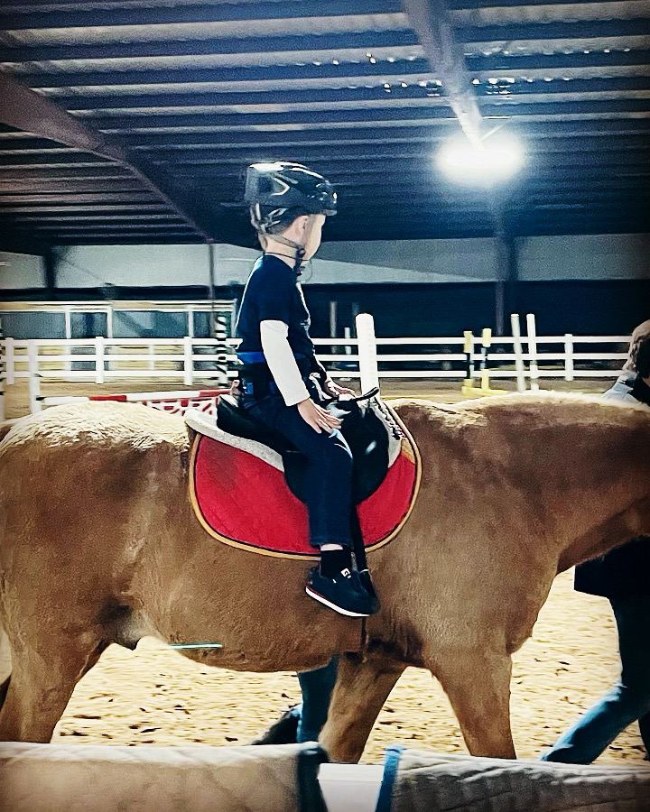 Un niño con casco monta un caballo alazán al paso en una arena techada, con una persona del equipo caminando a su lado.