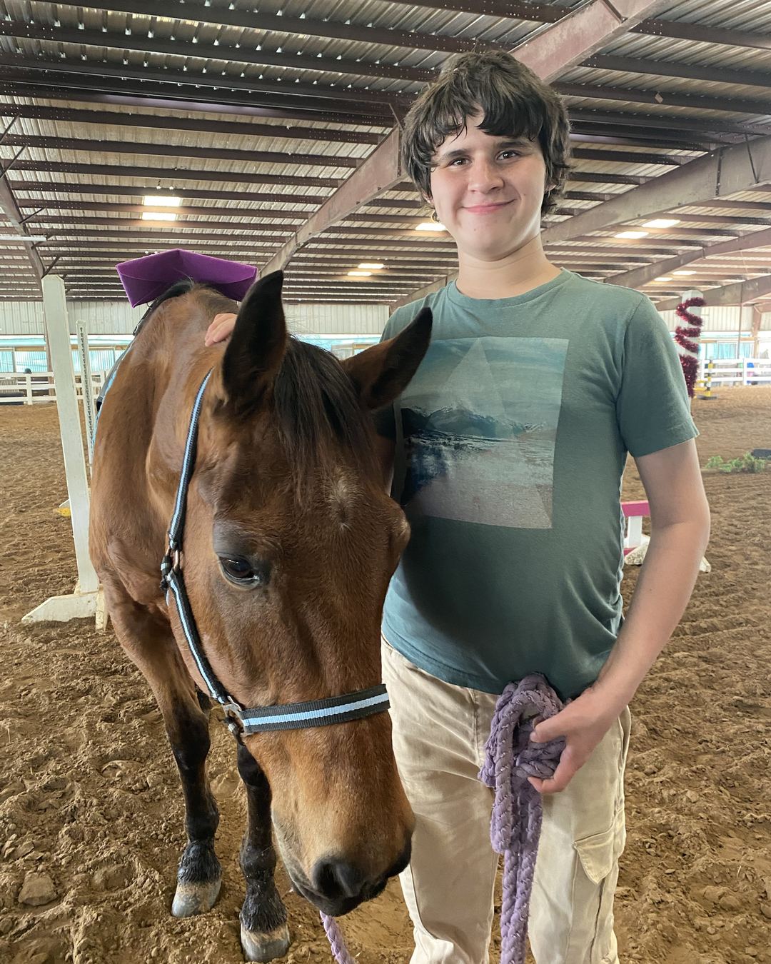 Un adolescente sonriente junto a un caballo bayo en una arena techada, sosteniendo la cuerda guía.